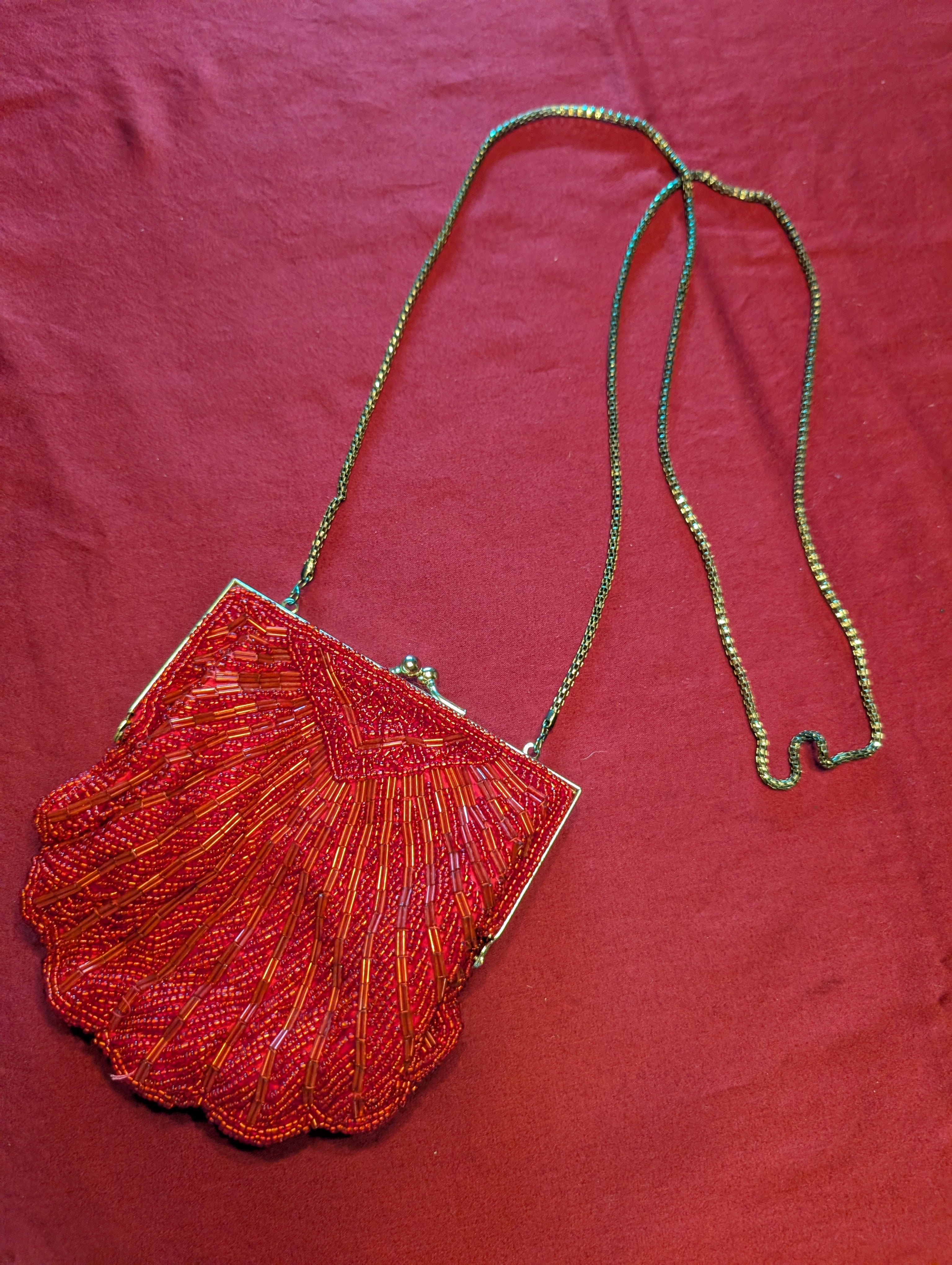 A vintage-style beaded clutch bag with a scalloped shell-shaped body, featuring red and orange beaded details, a silver-tone metal frame, and a thin metallic chain strap, displayed on a textured red fabric background.
