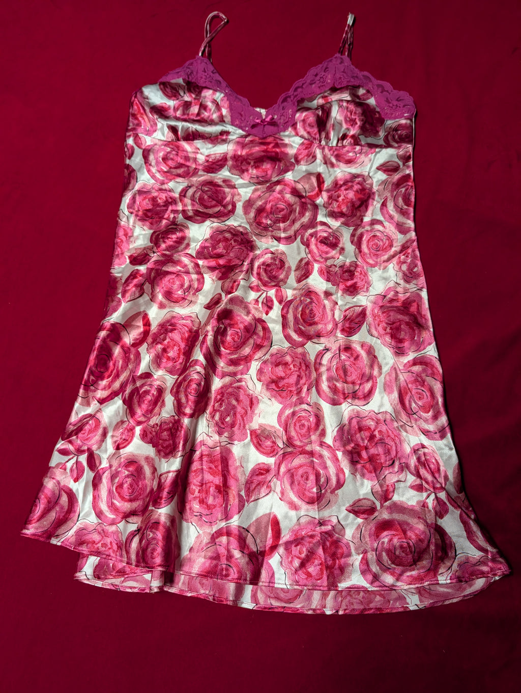 A sleeveless satin slip dress featuring a pink and red rose print on a white background, with a solid magenta lace-trimmed sweetheart neckline and spaghetti straps, laid flat on a dark red fabric background.