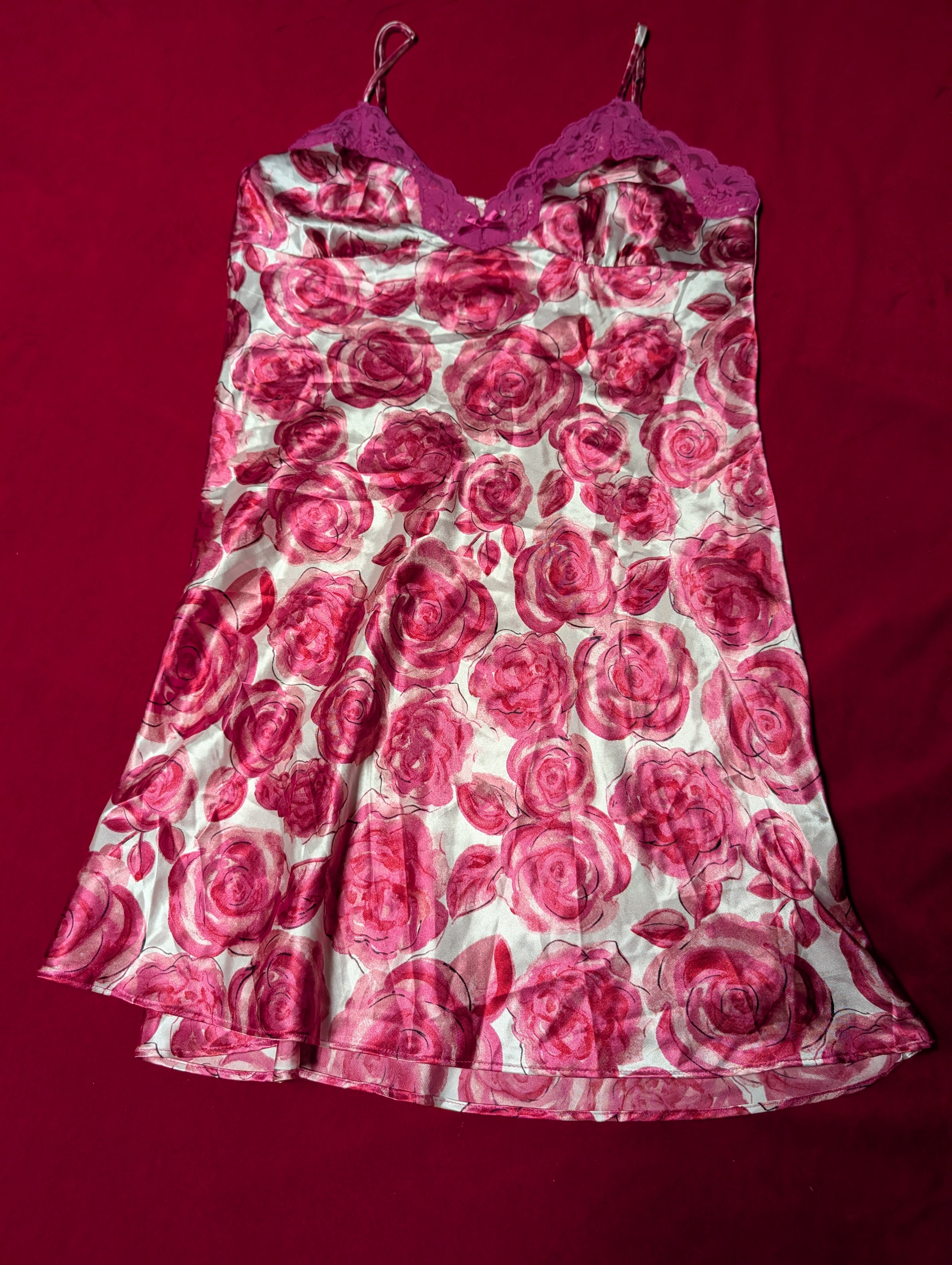 A sleeveless satin slip dress featuring a pink and red rose print on a white background, with a solid magenta lace-trimmed sweetheart neckline and spaghetti straps, laid flat on a dark red fabric background.